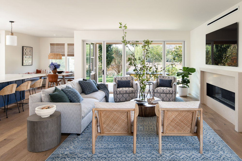 Open-concept living room layout showcasing a gray sofa, woven accent chairs, and patterned armchairs arranged around a wood coffee table, with a modern fireplace and large sliding glass doors leading to the backyard.