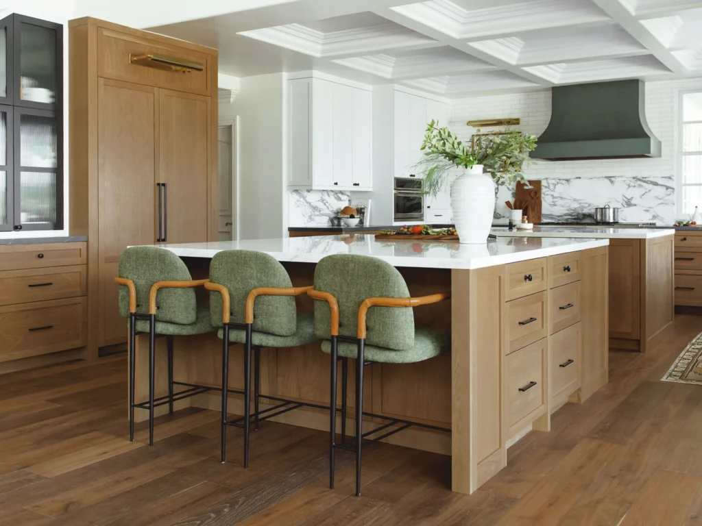 Warm modern kitchen with a large marble island, green upholstered bar stools, light wood cabinetry, and a coffered ceiling creating a designer focal point.