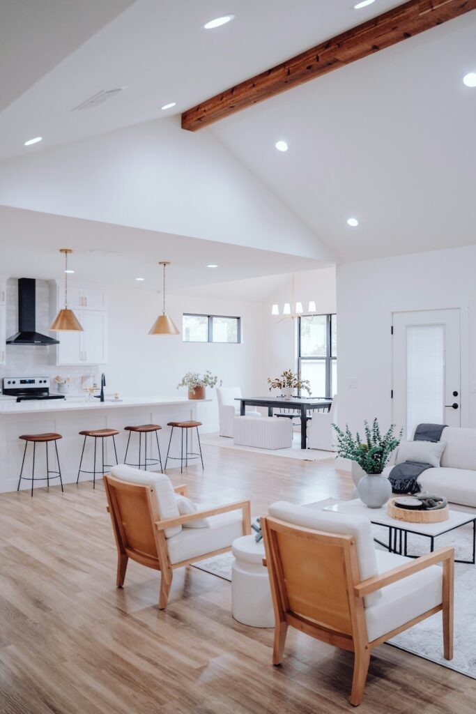 Bright open-concept living space with cohesive wood flooring, white walls, and light neutral furniture connecting the kitchen, dining, and living areas.