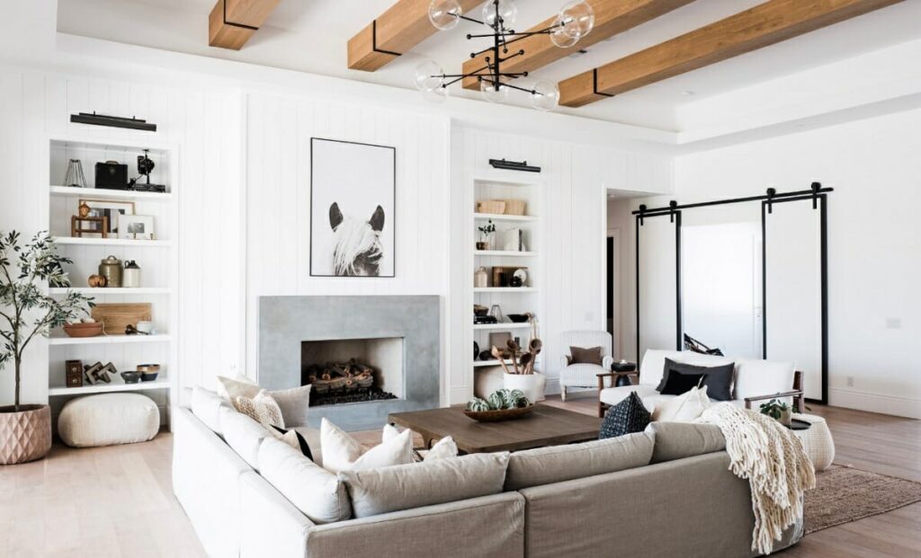 A bright modern farmhouse living room with white shiplap walls, exposed wood ceiling beams, and a large beige sectional facing a sleek concrete fireplace. Built-in shelves styled with decor flank the fireplace, while a modern black barn door and a sculptural chandelier complete the airy, cozy look.
