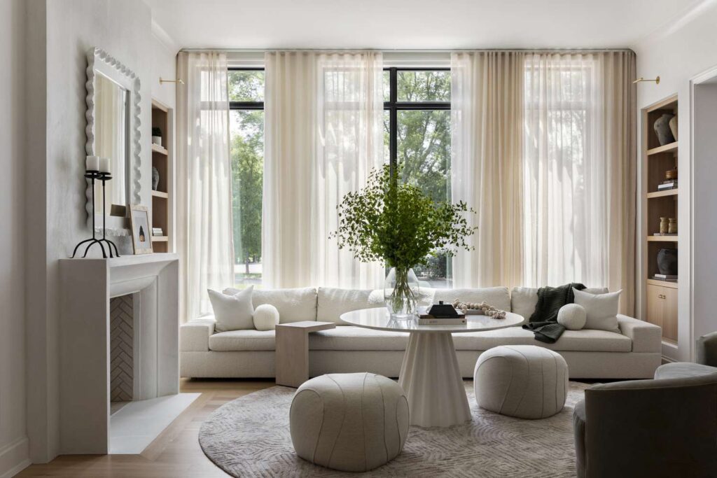 A bright living room with floor-to-ceiling windows, sheer curtains, a long white sofa, round ottomans, and a circular rug with a pedestal coffee table topped with a large vase of greenery.