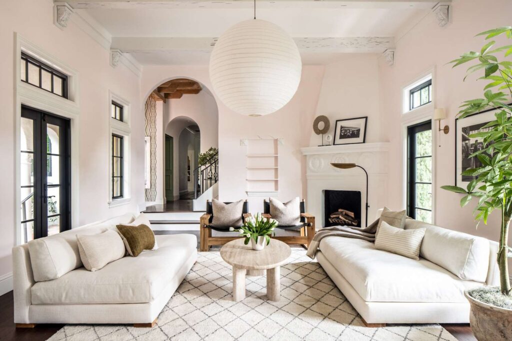 A soft, airy living room with twin white sofas facing each other, a round stone coffee table, arched doorways, black-framed windows, and layered neutral textiles.