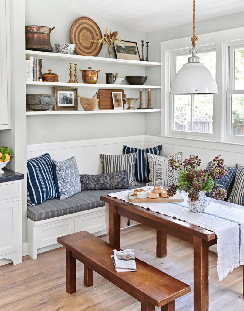 Kitchen banquette with cushioned bench seating and hidden storage, styled with layered pillows and open shelves displaying collected decor.