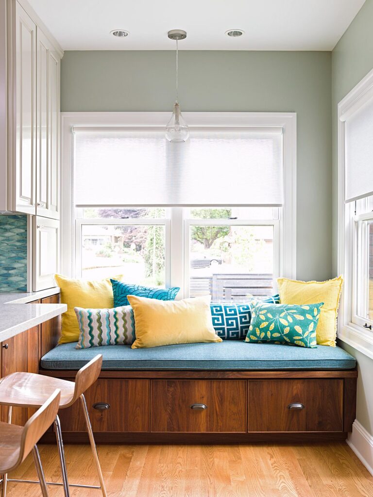 Window bench with built-in drawers and bright patterned pillows, doubling as seating and concealed storage in a compact kitchen space.