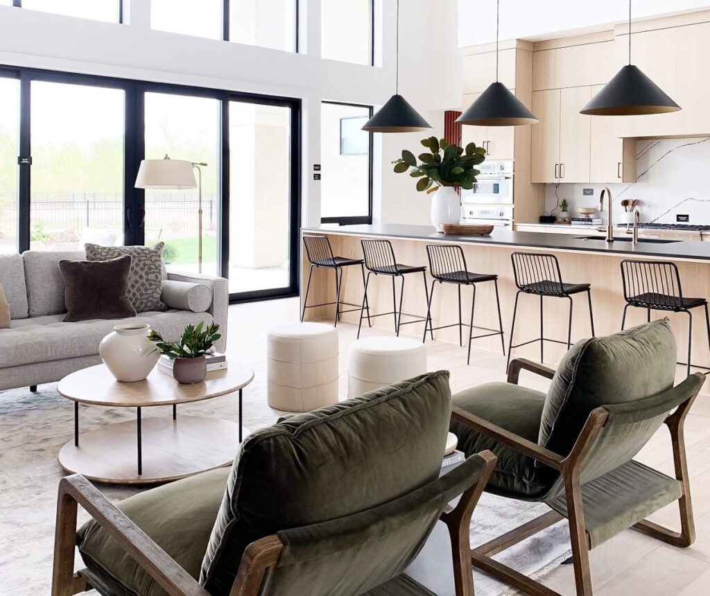 Bright open-concept living space with green lounge chairs, light wood kitchen cabinets, black pendant lights, and a modern living-to-kitchen layout.