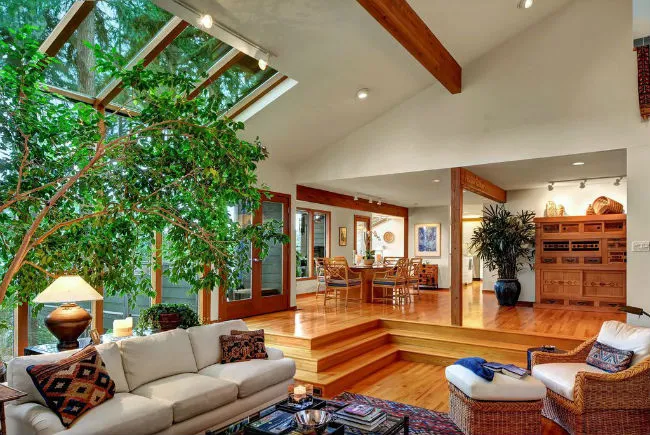 Split-level open-concept home with a sunken living room, large skylight windows, warm wood floors, and natural greenery separating the dining and kitchen areas.