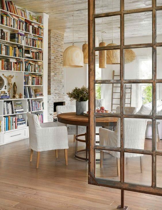 Open-concept dining area with built-in bookshelves, rustic glass partition, and layered natural textures connecting to a bright living space