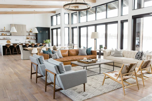 Open-concept living room with large windows, a leather sofa, modern gray chairs, and a connected kitchen and dining area showing a zoned layout.