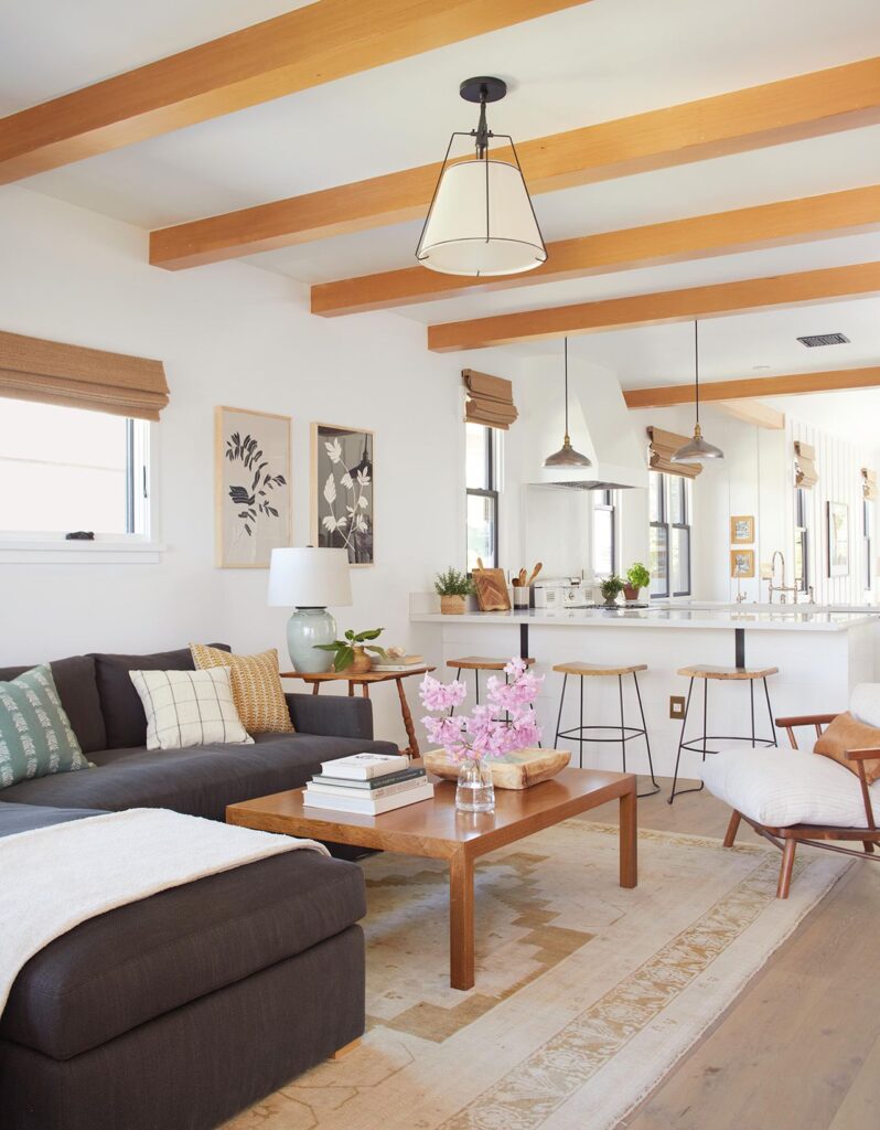 Warm open-concept living room with wood ceiling beams, a dark sectional, and a connected kitchen bar area styled with natural textures and soft zoning.